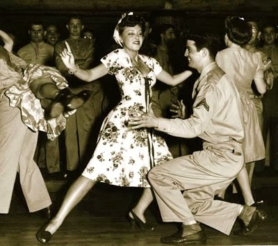 1940s vintage photo of people swing dancing in the 1940s. The woman is wearing a floral 1940s dress and matching hair turban / hair scarf. 