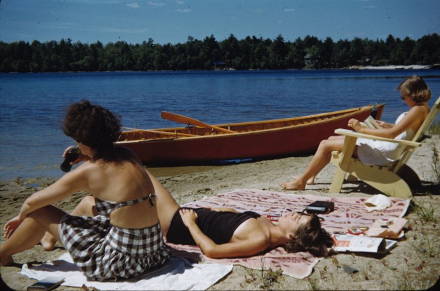 1950s vintage photo of 3 women in 1950s swimsuits sitting on a beach by a boat. 